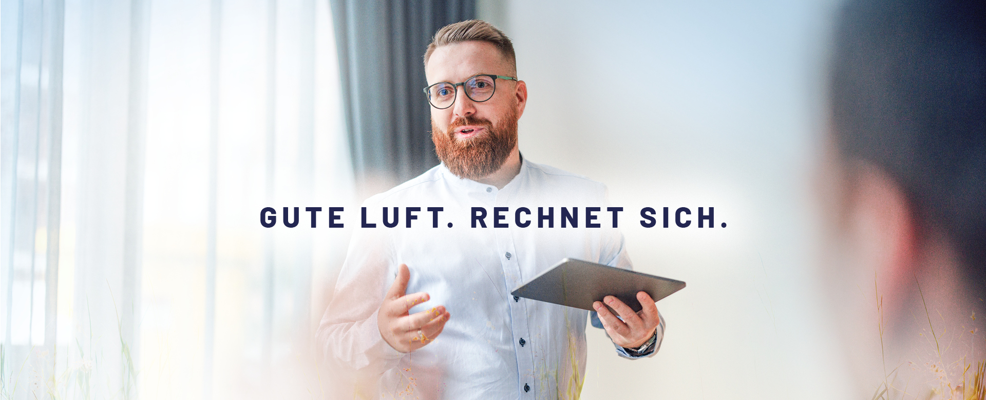 Foto mit dem Text „Gute Luft. Rechnet sich“. Im Hintergrund ist ein Mann mittleren Alters zu sehen, der ein Tablet in der Hand hält und Professionalität ausstrahlt.
